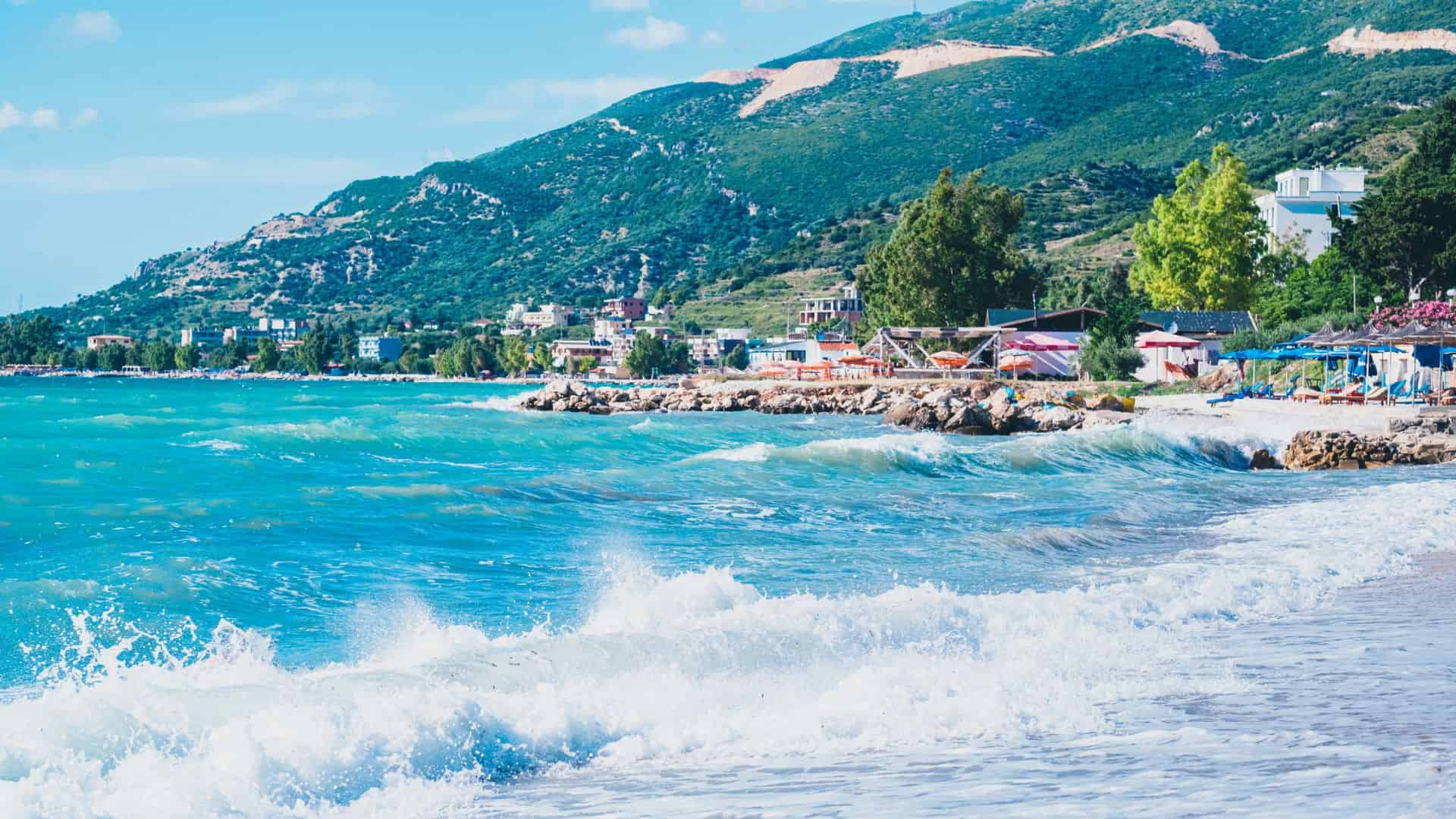 Waves crashing on a turquoise beach with colorful umbrellas and green hills in the background.