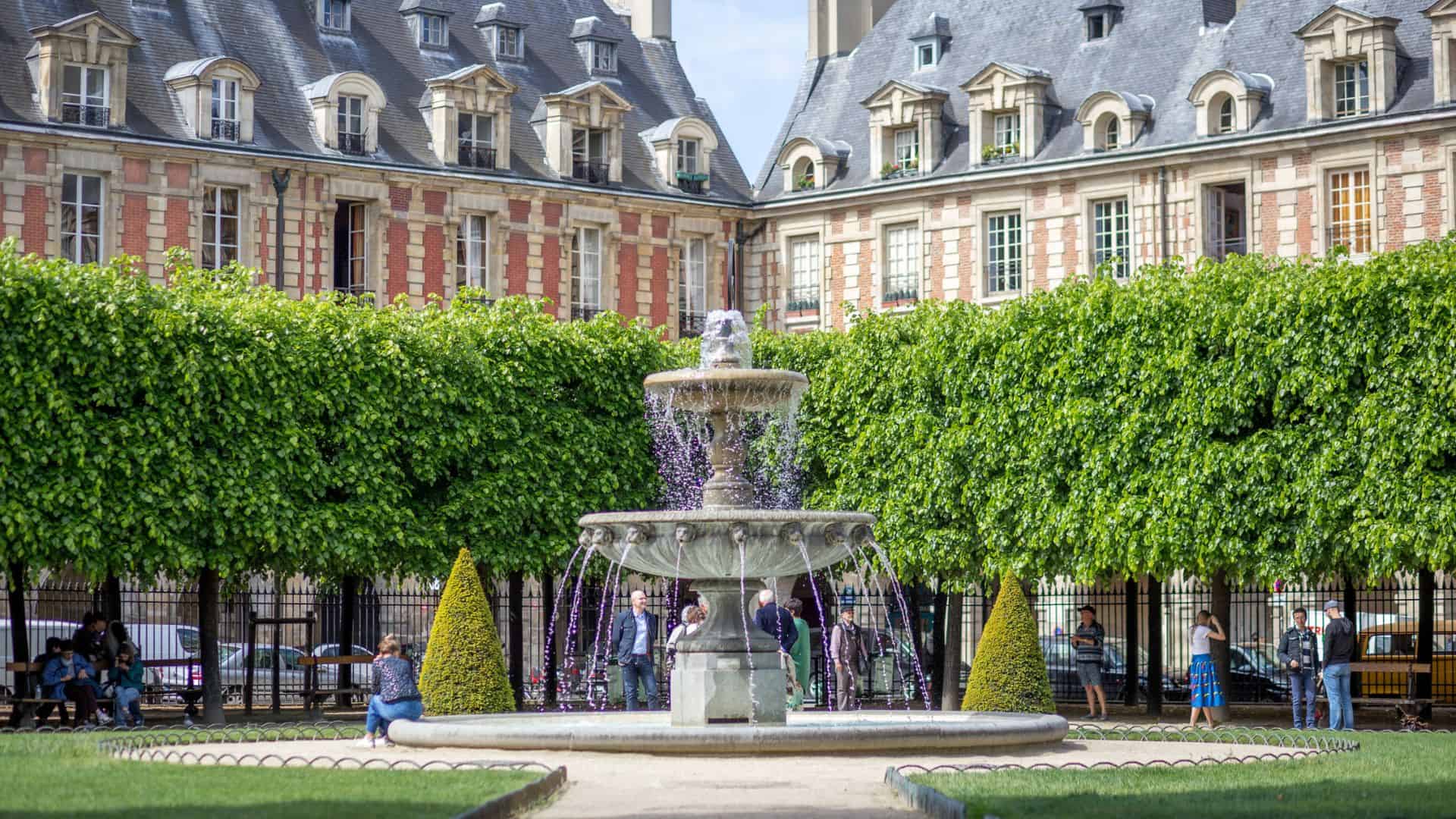 A stone fountain surrounded by trimmed trees in a courtyard with historic buildings in the background.
