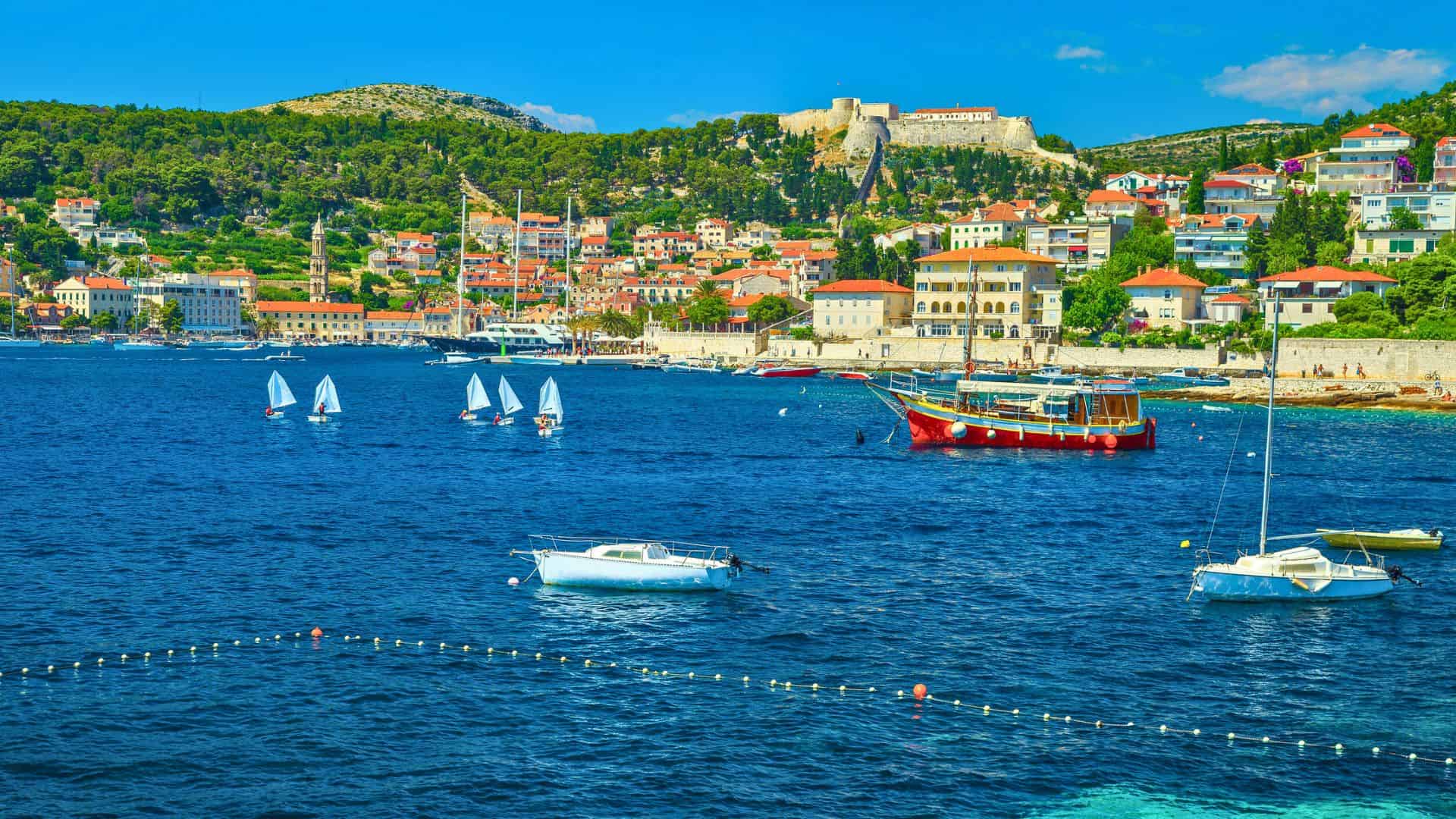 Boats and sailboats on blue water with a coastal town and hillside fortress in the background under a clear sky.