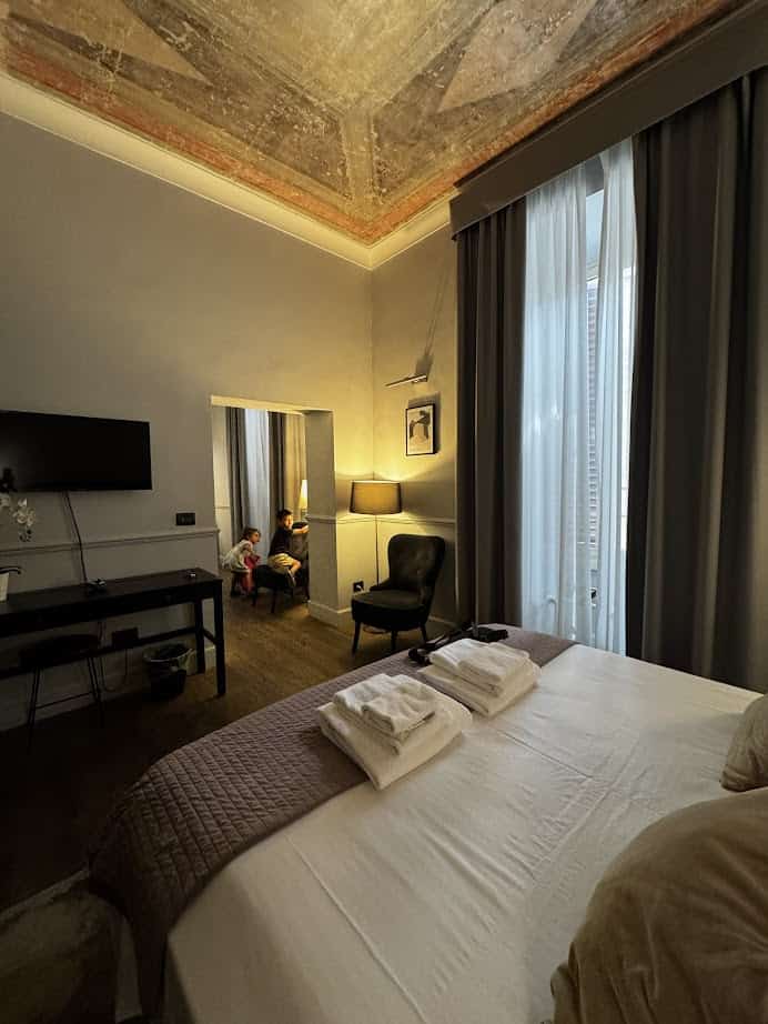 Elegant hotel room with a large bed, TV, high ceiling, and two children sitting in a cozy reading nook—perfect for families planning their Rome with Kids adventure.