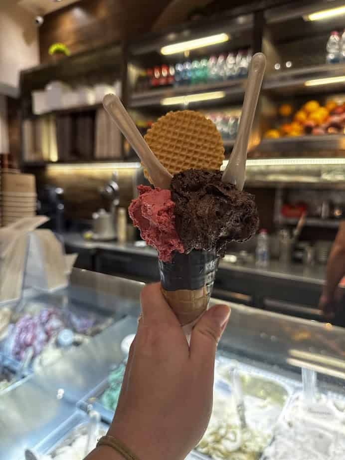 A hand holds an ice cream cone with two scoops, a waffle, and two spoons inside a gelato shop—a sweet stop for anyone searching where to eat in Rome.