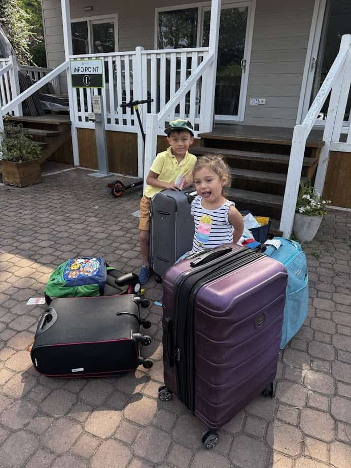 Two children stand outside with several large suitcases and backpacks in front of a building with stairs, ready to begin their adventure at Parco della Gallinara.