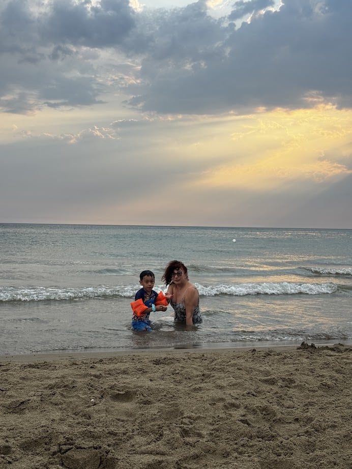 Child and adult playing in shallow sea water at sunset, with waves and a cloudy sky in the background, reminiscent of tranquil evenings near Parco della Gallinara.