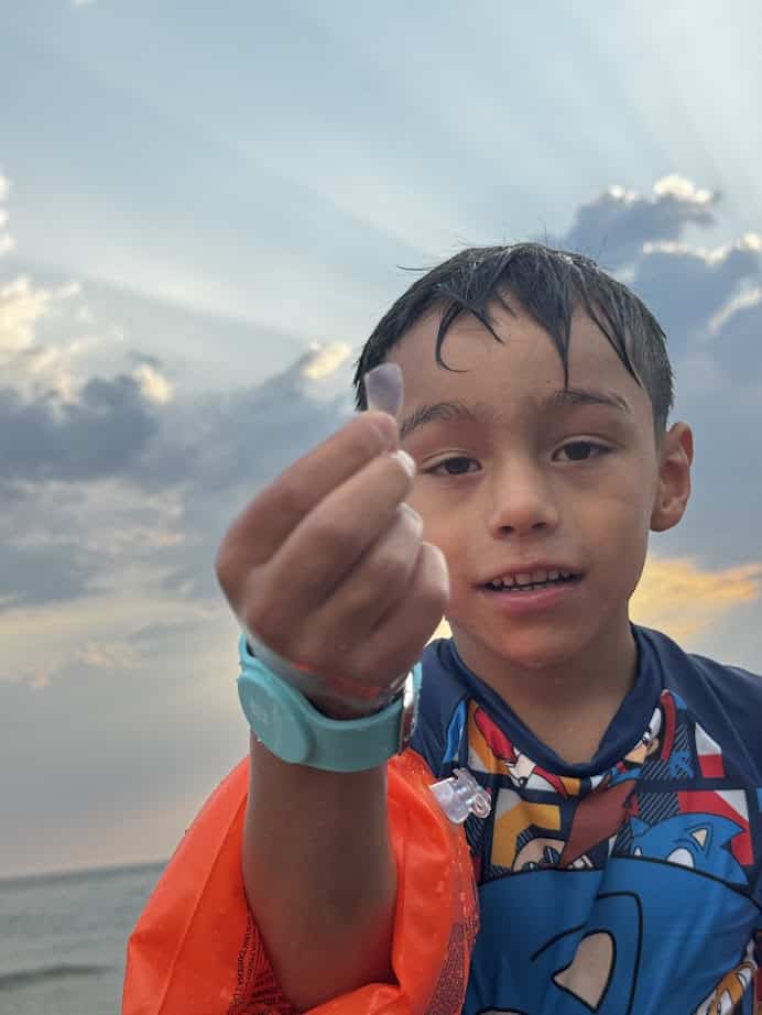A boy at the beach in Parco della Gallinara holds up a small object, smiling, with dramatic clouds and sunlight in the background.