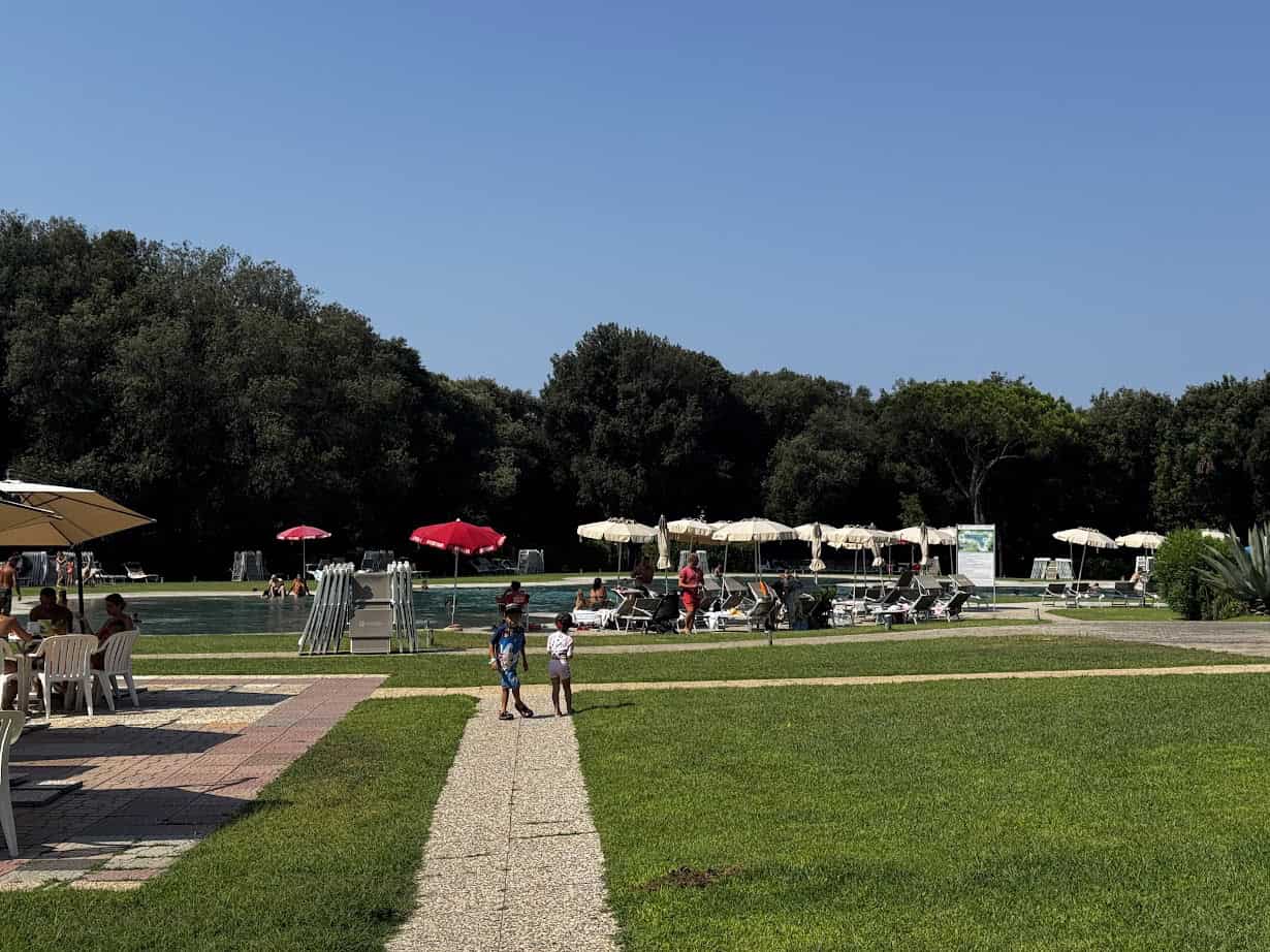 People relaxing by a pool with umbrellas at Parco della Gallinara, trees in the background, and children walking on a path in front.