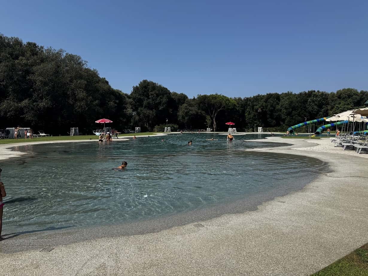 People swim in a large outdoor pool at Parco della Gallinara, surrounded by trees, lounge chairs, and umbrellas under a clear sky.