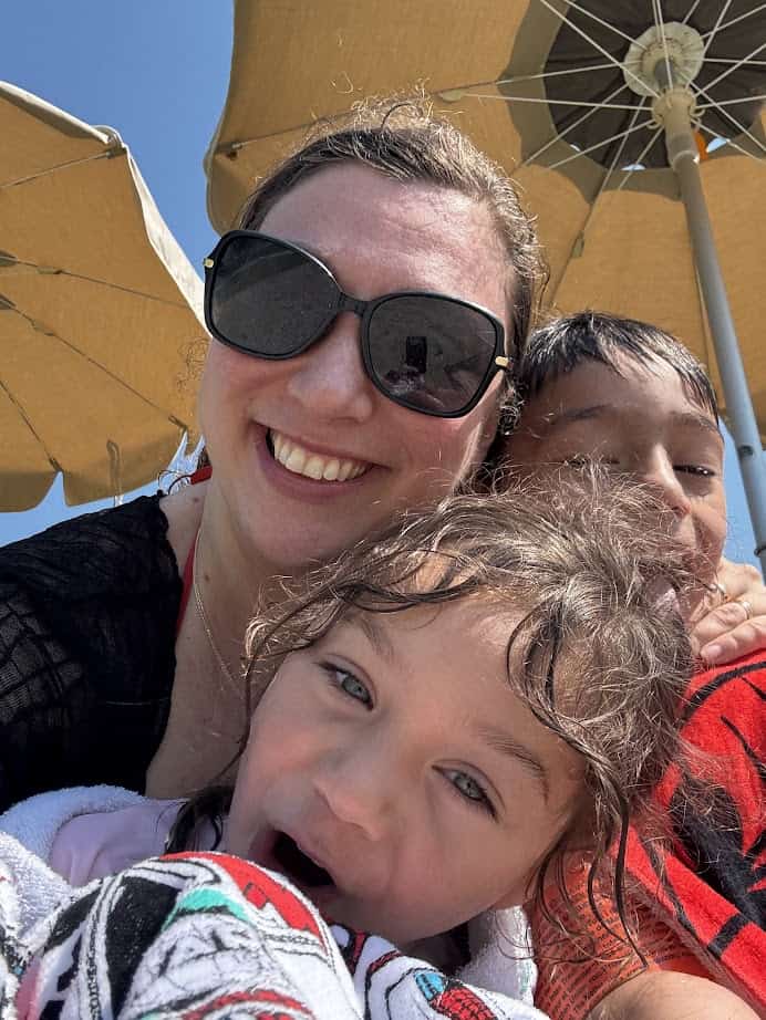 Woman in sunglasses smiling with two children under large beige beach umbrellas at Parco della Gallinara on a sunny day.