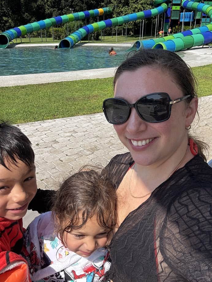 A woman and two children smile at Parco della Gallinara, with water slides and a sparkling pool in the background.