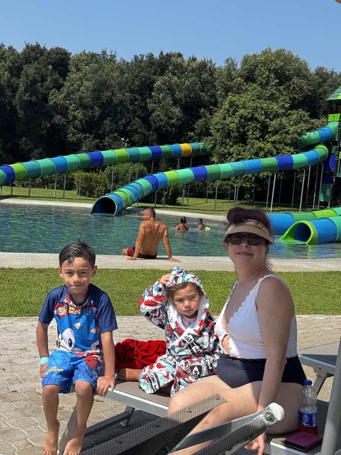 A woman and two children sit poolside at Parco della Gallinara, a water park with colorful slides and green trees in the background.