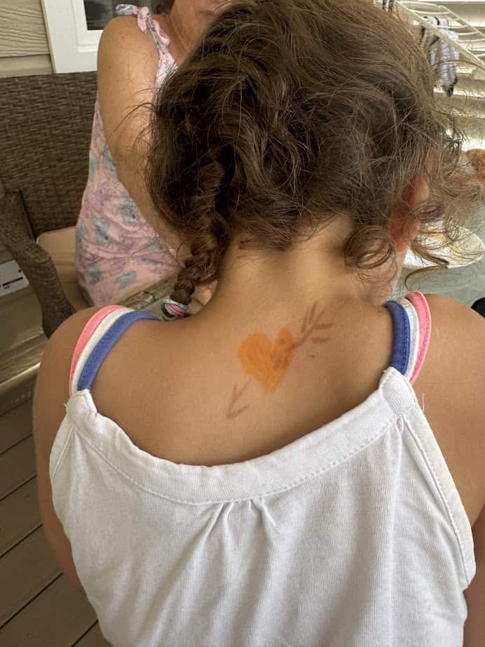 Young girl with braided hair and a heart drawing on her upper back, wearing a white tank top, enjoys the sunshine at Parco della Gallinara.