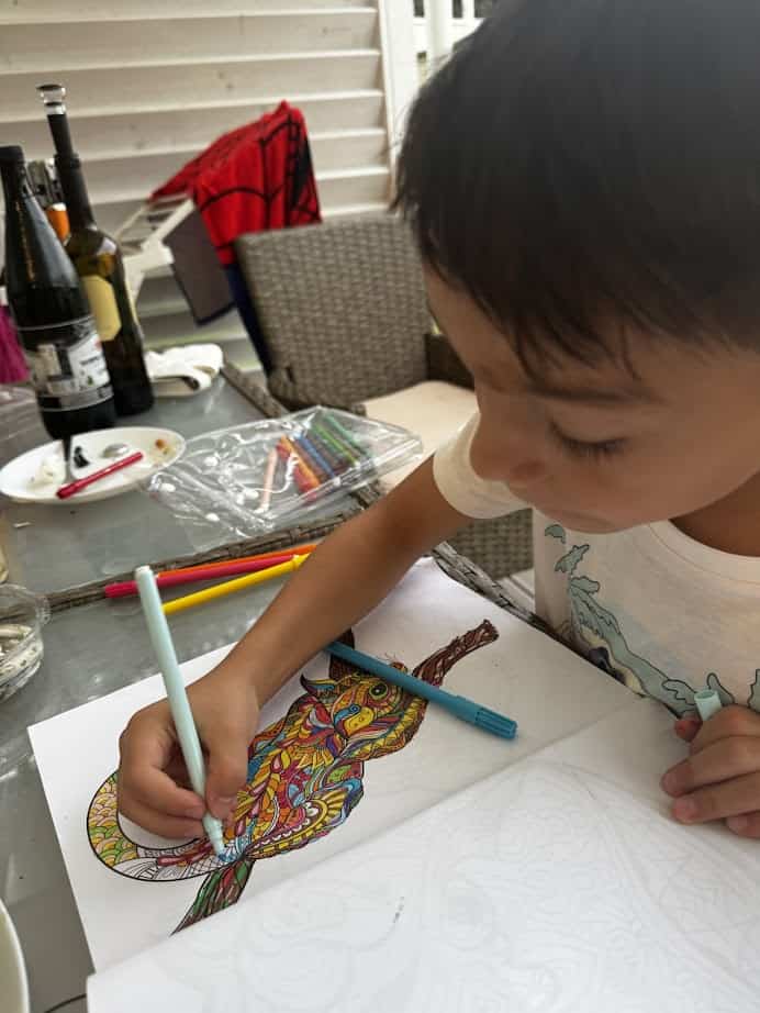 A child colors a detailed bird drawing at a table in Parco della Gallinara, with markers, bottles, and dishes in the background.