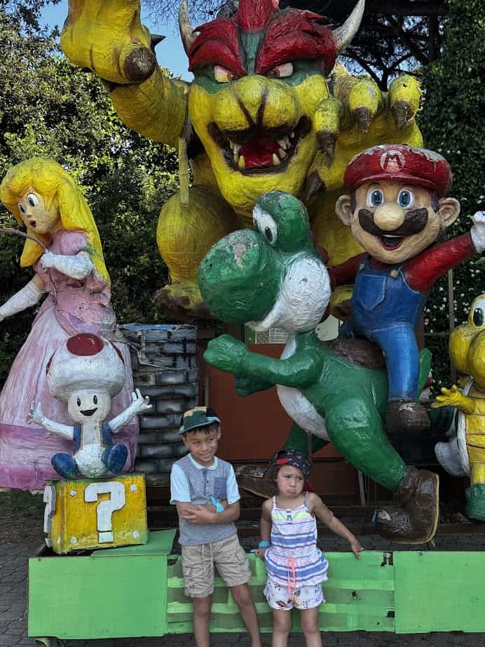 Two children pose in front of large statues of Mario characters, including Mario, Yoshi, Bowser, and Princess Peach, at parco della gallinara.