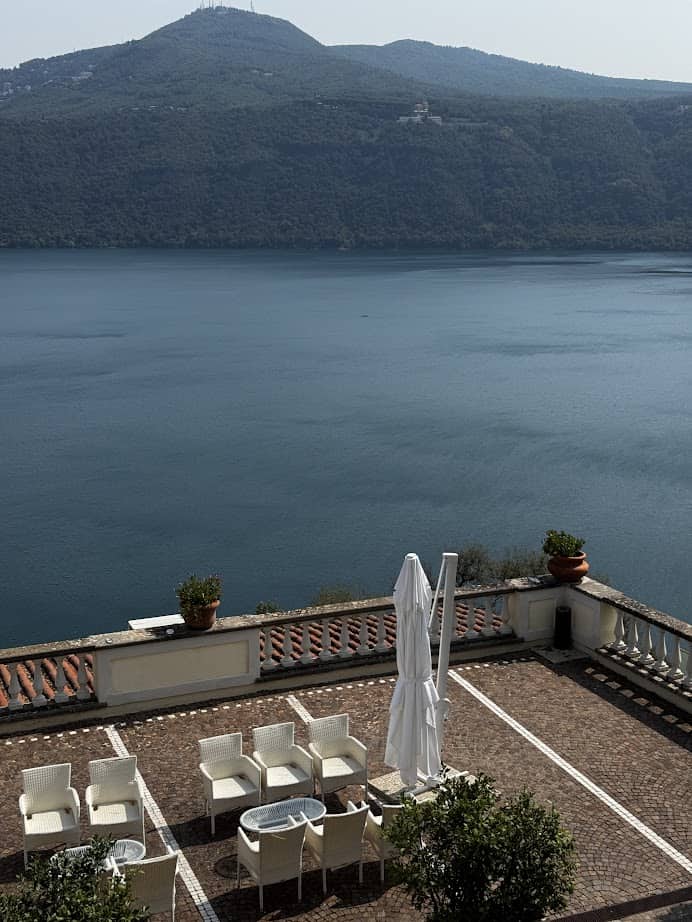 Outdoor patio with white chairs and tables overlooking a calm lake and green hills in the background—an ideal spot to relax during your 4 days in Castel Gandolfo.