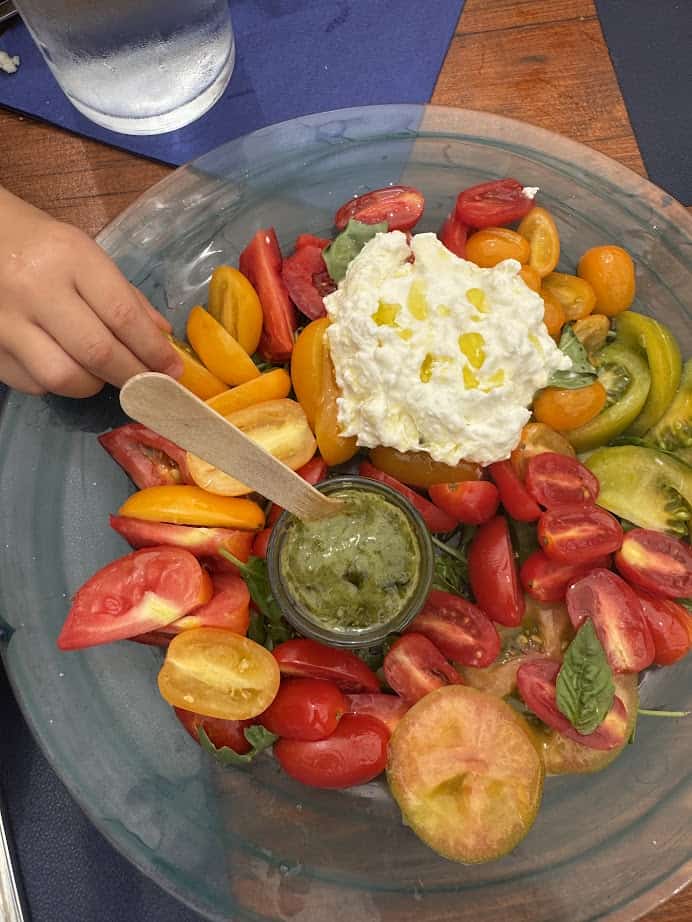 A child’s hand reaches for colorful tomatoes, burrata cheese, and pesto on a clear plate—capturing the simple delights found during 4 days in Castel Gandolfo.