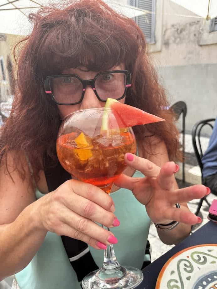 Woman with curly hair and glasses holding a cocktail garnished with a watermelon slice, enjoying the sunshine outdoors during her 4 days in Castel Gandolfo.