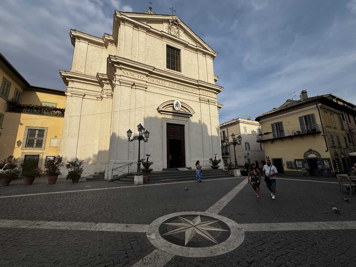 Visitors enjoying 4 days in Castel Gandolfo walk in front of a large beige church, with a compass rose set into the cobblestone plaza in the foreground.