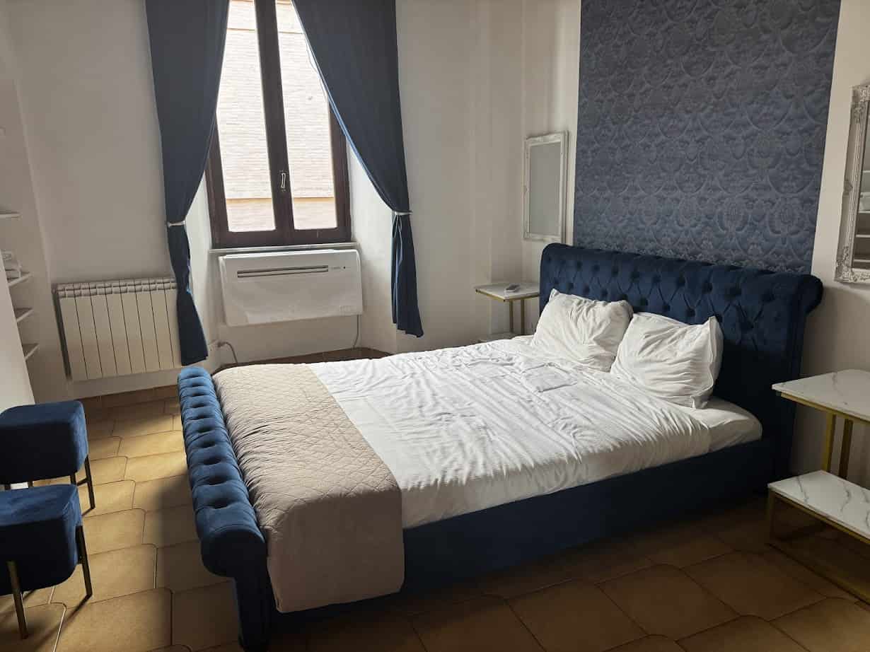 A neatly made bed with a blue velvet frame in a bright room with large window and elegant decor—perfect for relaxing after a day exploring Rome with kids.