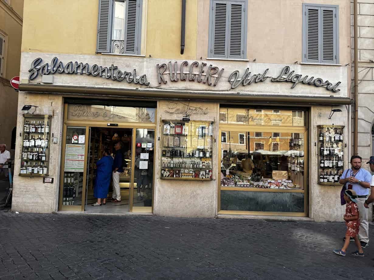 Storefront of Salsamenteria Ruggeri with bottles in the window and people walking outside on a cobblestone street, a highlight for anyone exploring the best food tours in Rome.