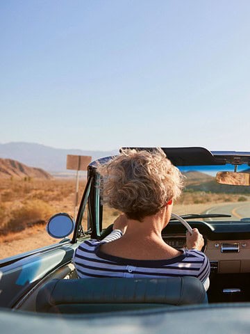 Two people in a convertible driving through a desert; one has arms raised joyfully under a clear blue sky.