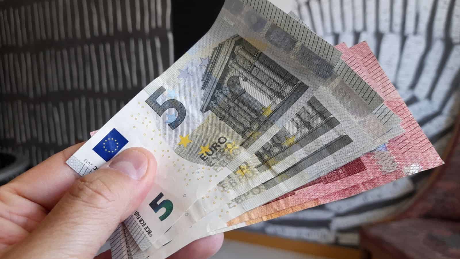 A hand holding 5, 10, and 20 euro banknotes, fanned out against a patterned background.