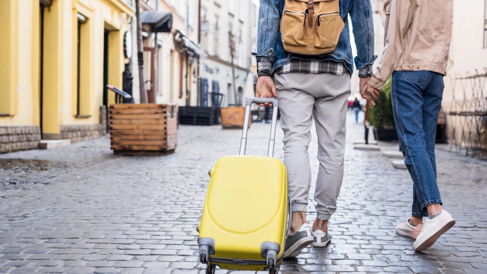 Two people holding hands walk down a cobblestone street, one pulling a yellow suitcase behind them.