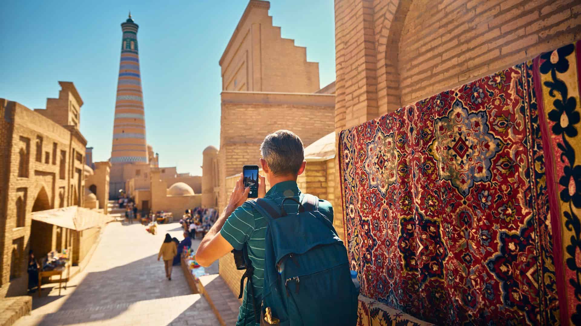 A man with a backpack photographs a tall minaret along a street lined with carpets in a historic city.