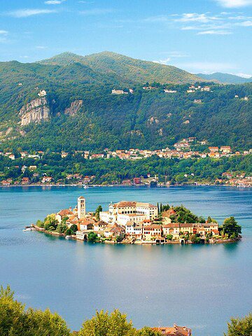 A small island with buildings sits in a blue lake, surrounded by green hills and distant mountains under a blue sky.