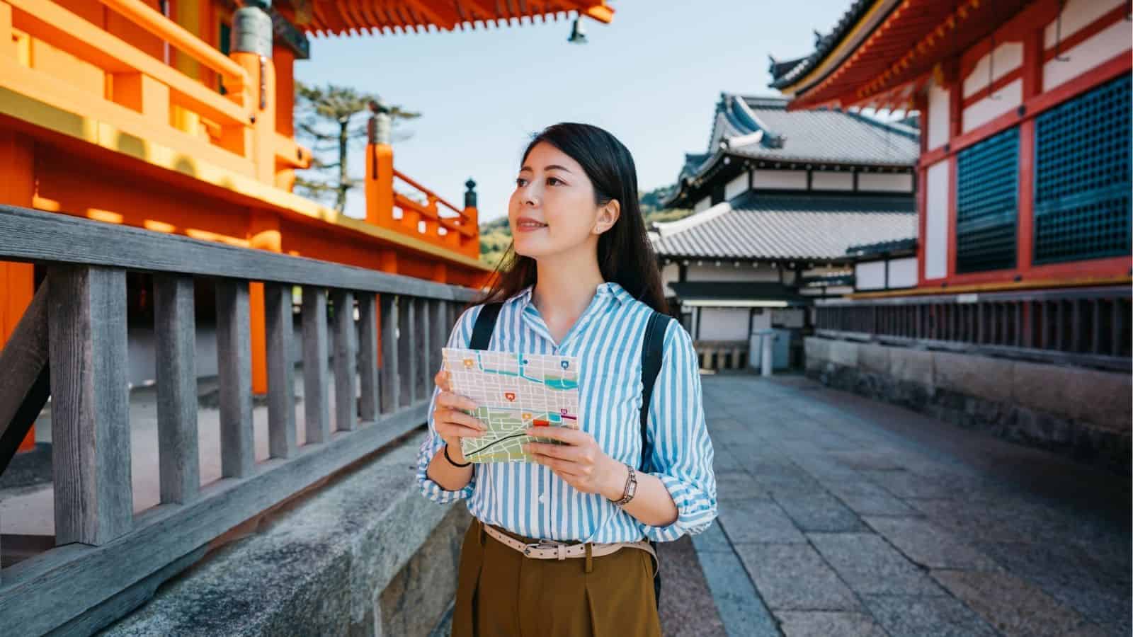 Woman holding a map, exploring traditional Japanese buildings on a sunny day, immersing herself in local customs and the charm of daily travel during her Asia travel adventure.