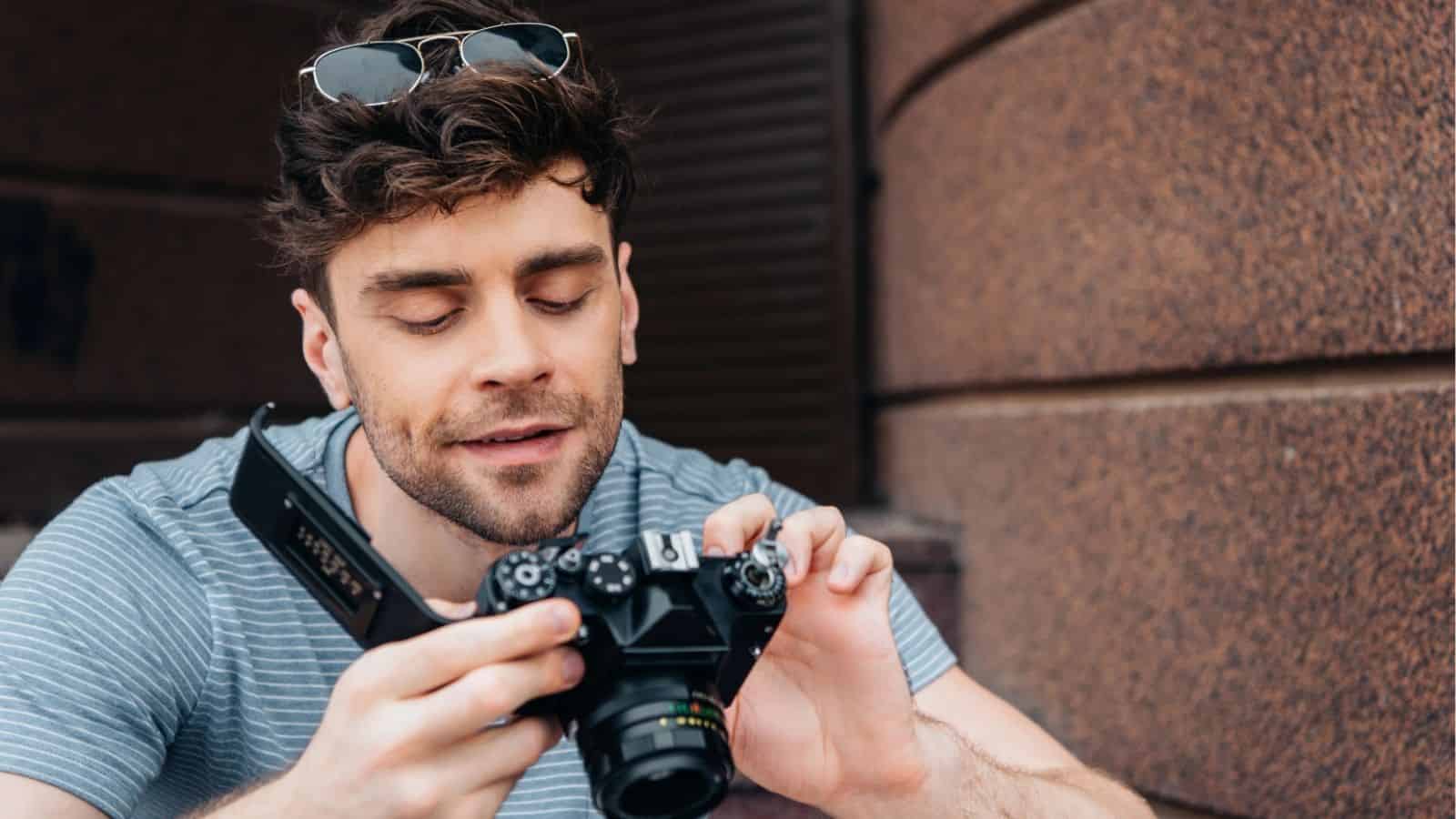 Man in a striped shirt looking down and adjusting settings on a black camera, with sunglasses on his head.