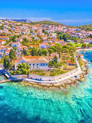 Aerial view of a coastal town with colorful buildings, clear blue water, and many boats anchored near shore.