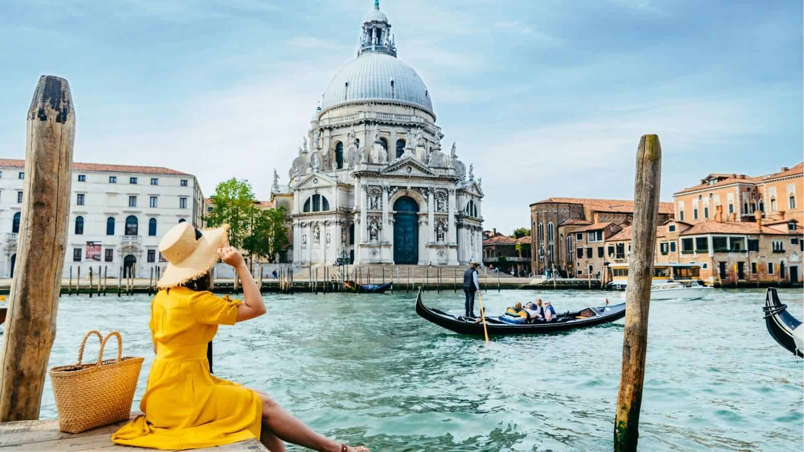 A woman in a yellow dress sits by a canal in Venice, watching a gondola near a large domed church.