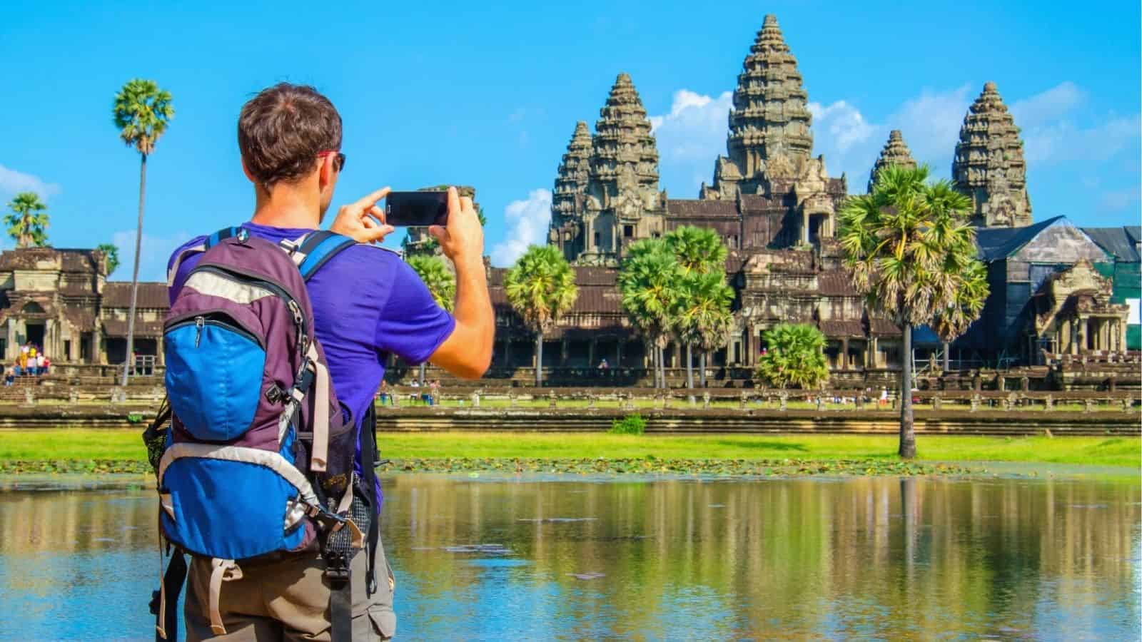 A tourist with a backpack takes a photo of the sacred site Angkor Wat temple by a pond on a sunny day.