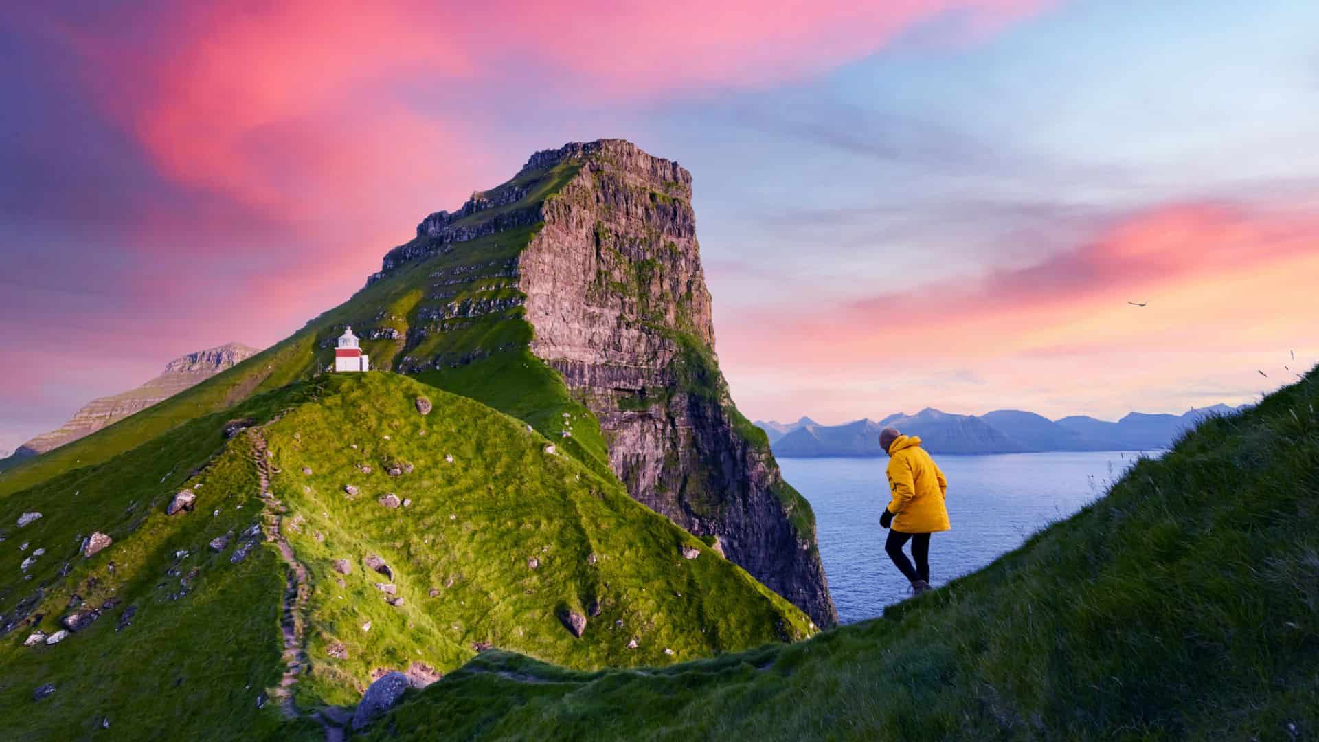Person in yellow jacket hikes grassy cliff toward small lighthouse, with pink sunset sky and sea in background.