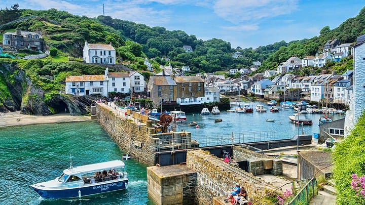 Picturesque coastal village with boats in the harbor, stone buildings, and green hills under a blue sky.