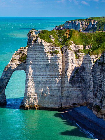 White chalk cliffs and a natural arch rise above turquoise sea at Étretat, Normandy, under a clear blue sky.