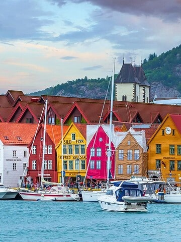 Colorful wooden buildings and boats along the waterfront in Bryggen, Bergen, Norway, with hills in the background.