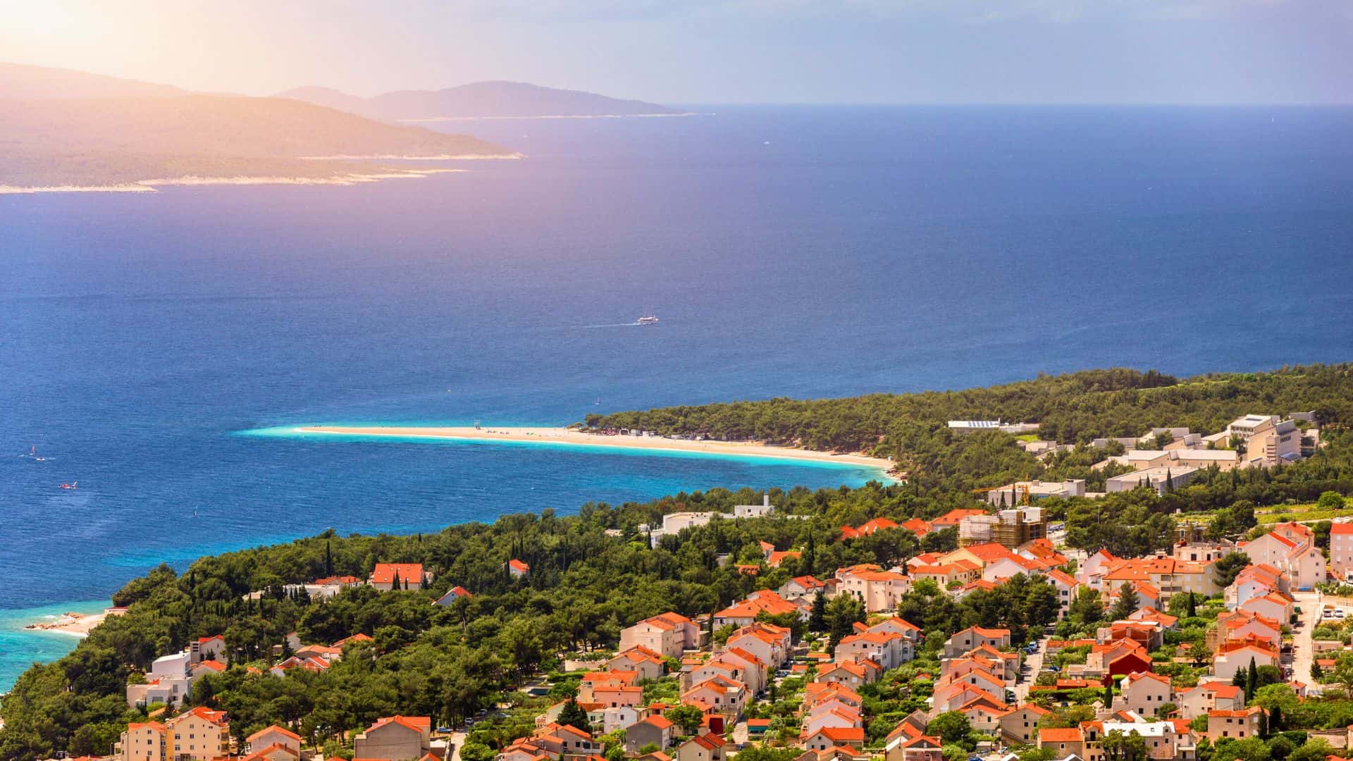 A coastal town with red-roofed houses overlooks a forest and a sandy beach by the blue sea under a sunny sky.