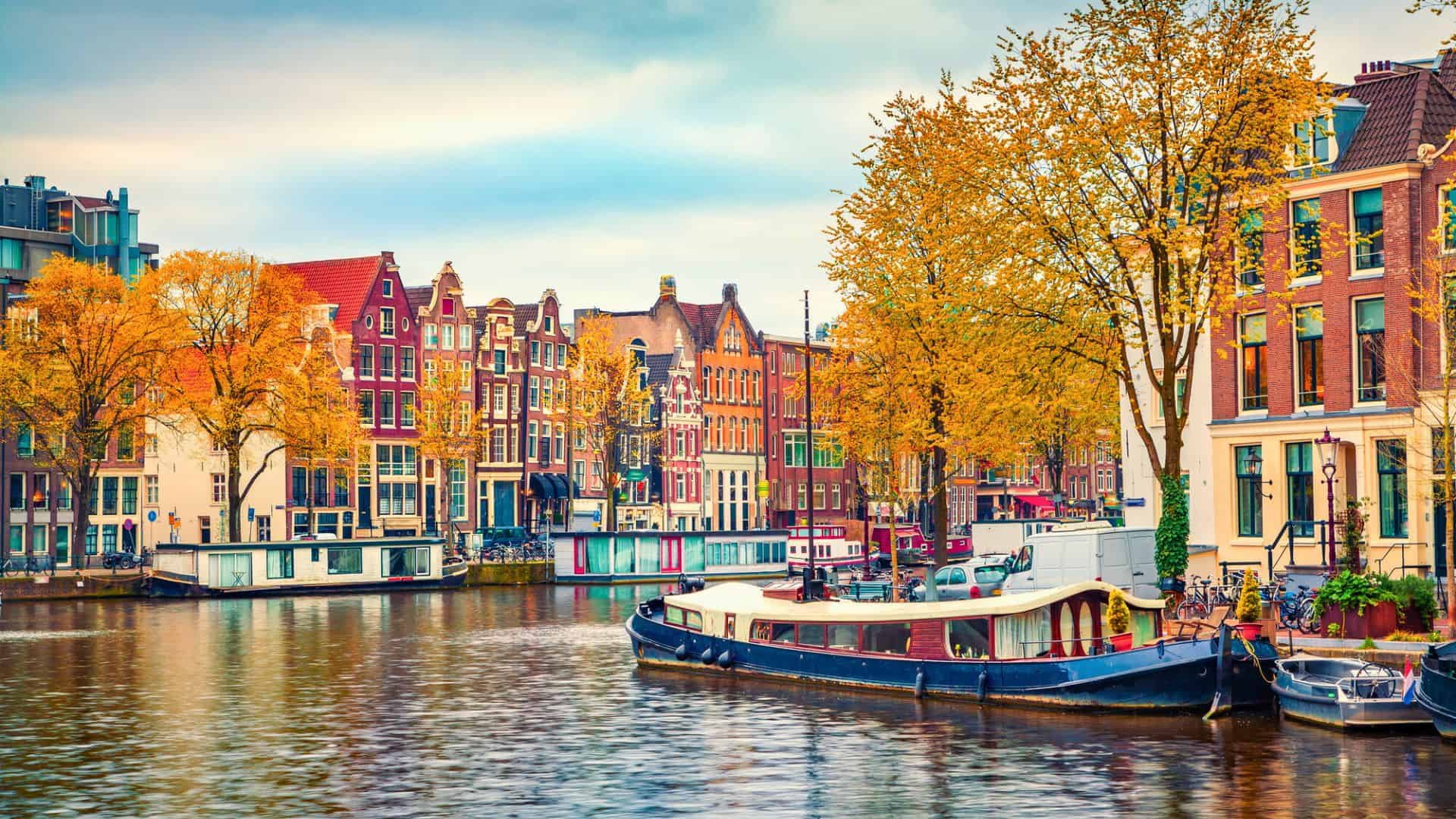 Colorful canal houses, boats, and autumn trees line a waterway in Amsterdam under a cloudy sky.