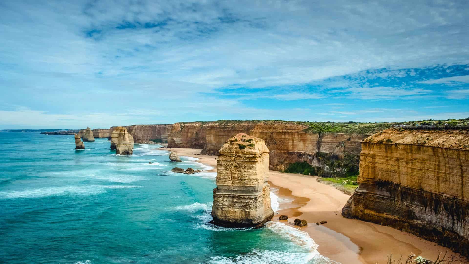 Rocky limestone stacks rise from the ocean along a sandy coastline under a partly cloudy sky.