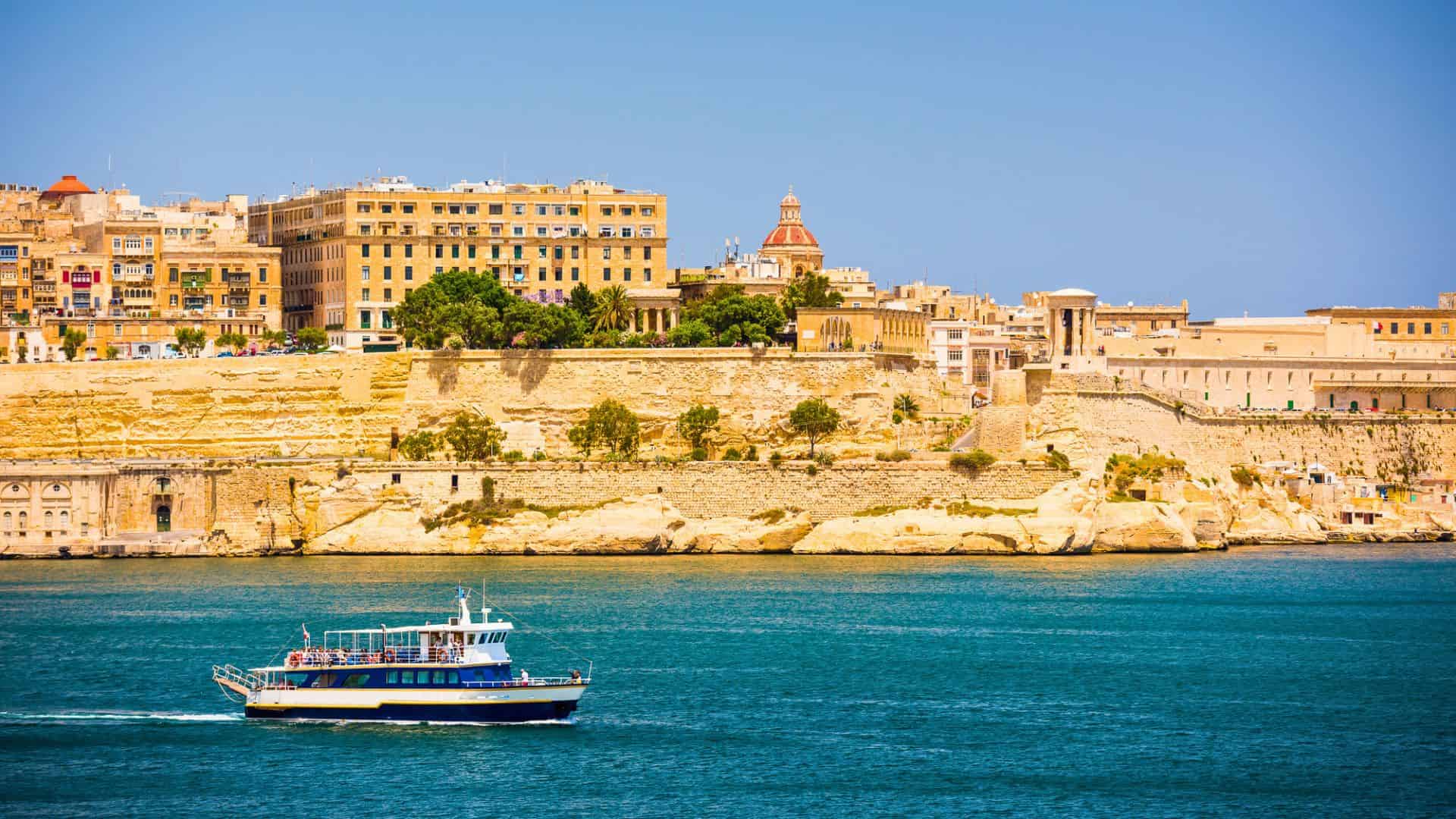 A blue and white boat sails on turquoise water near historic buildings atop golden cliffs.