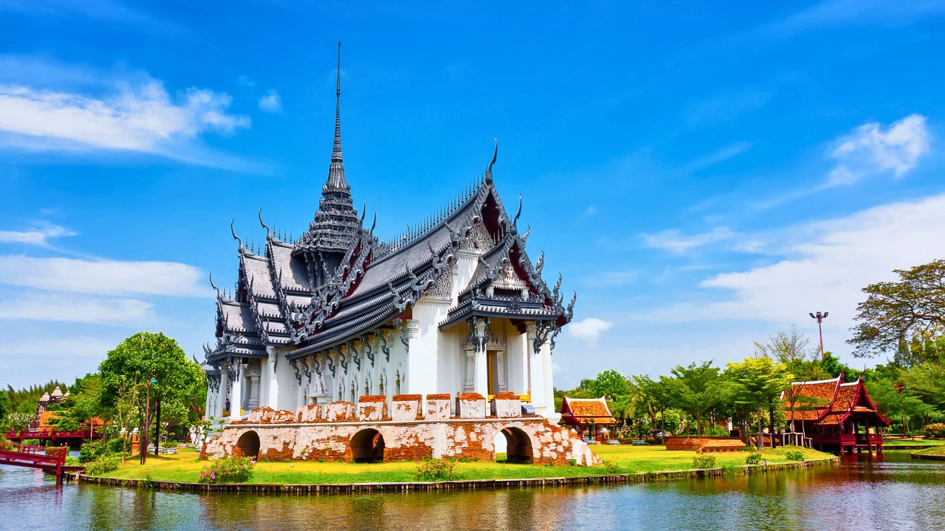 Traditional Thai temple with ornate roof by a pond, surrounded by greenery under a clear blue sky.