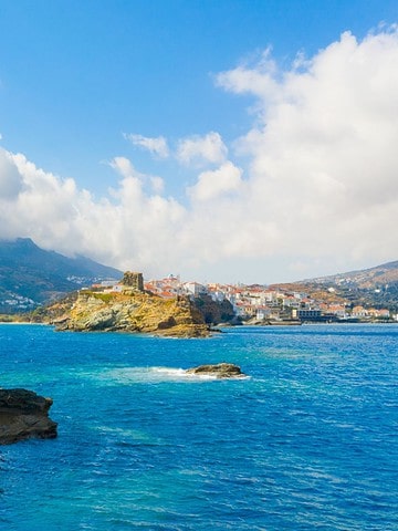 A stone lighthouse stands on a rocky islet in blue sea, with a coastal town and mountains in the background.