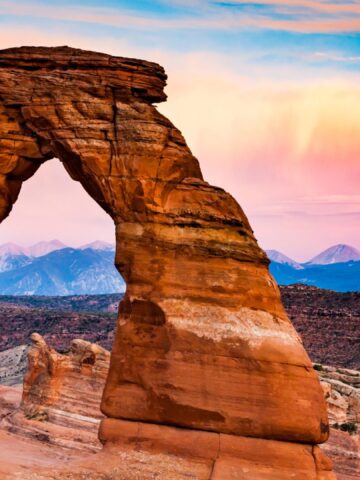 Delicate Arch at sunset with colorful sky and distant mountains in Arches National Park, Utah.