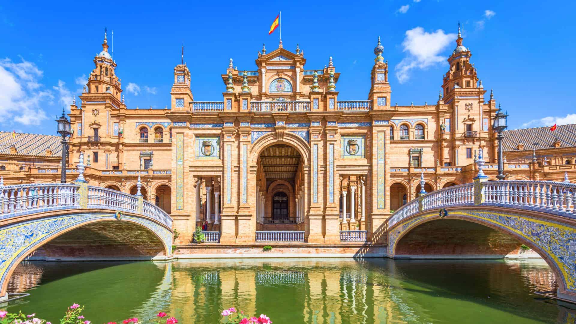 Ornate Spanish building with towers, bridges, and a canal beneath a bright blue sky, with a Spanish flag on top.