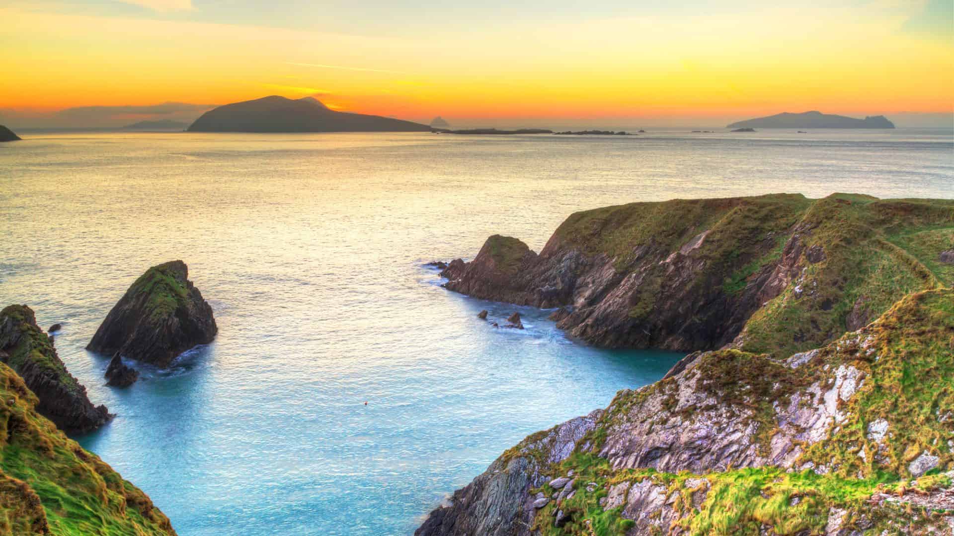 Rocky coastal cliffs and blue-green sea at sunset, with distant islands under an orange sky.