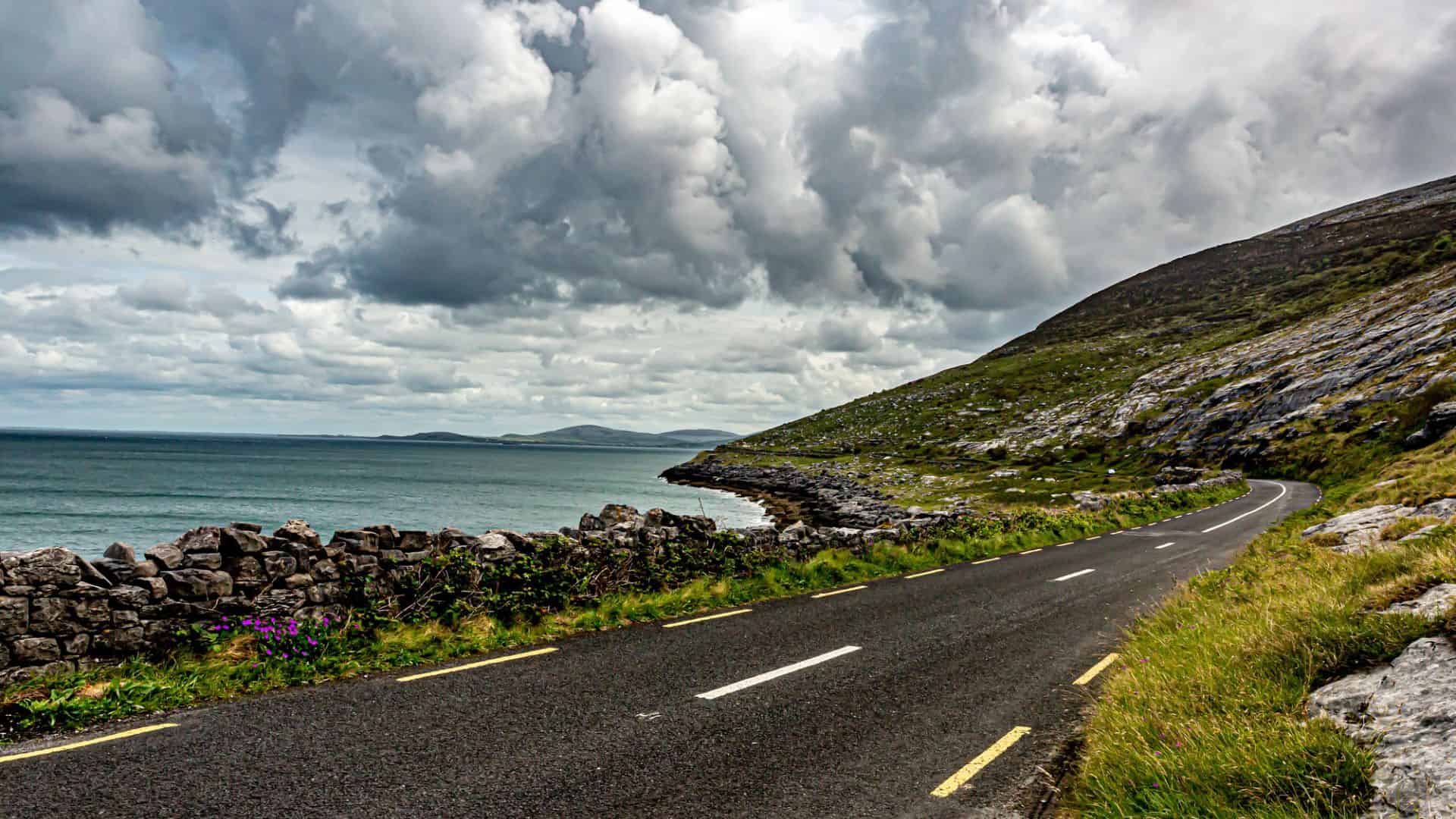 A winding coastal road runs by the sea under dramatic, cloudy skies with rocky hills and green grass.