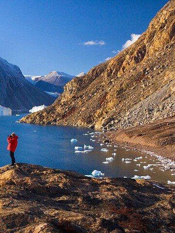 Person in a red jacket stands on rocky shore, photographing icebergs on a blue lake with snowy mountains behind.