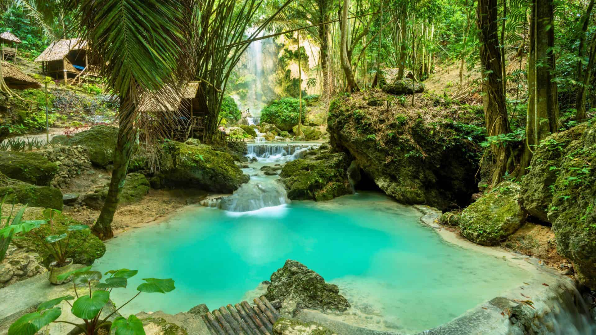 Turquoise waterfall pool surrounded by lush green trees and small wooden huts in a tropical jungle.