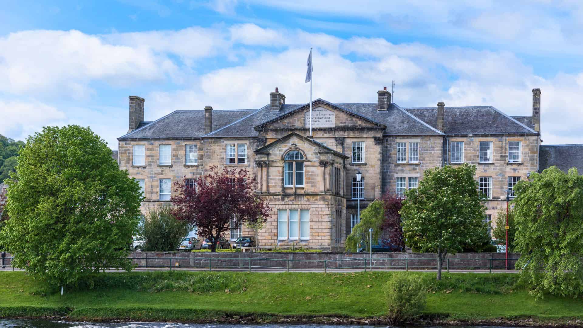 Historic stone building with a central flagpole, surrounded by trees and a river in the foreground.