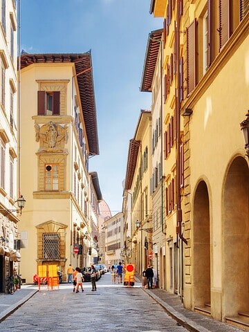 Narrow cobblestone street lined with historic buildings, pedestrians, and warm sunlight in a European city.
